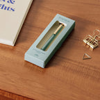 Green pen in a box labeled 'PAPIER' on a wooden surface with office supplies.