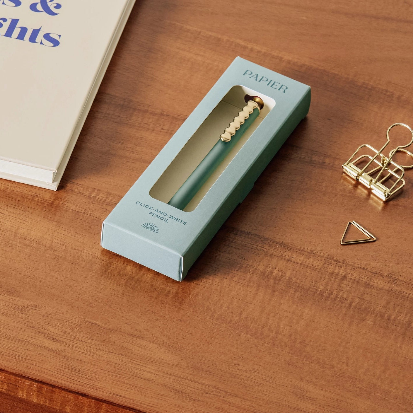 Green pen in a box labeled 'PAPIER' on a wooden surface with office supplies.